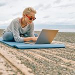 Positive well-fit blond woman in sportswear, seated in lotus pose, using laptop at the beach. Freelance working, wellbeing concept.