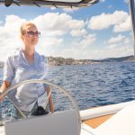 Attractive blond female skipper navigating the fancy catamaran sailboat on sunny summer day on calm blue sea water. Luxury summer adventure, active nautical vacation.